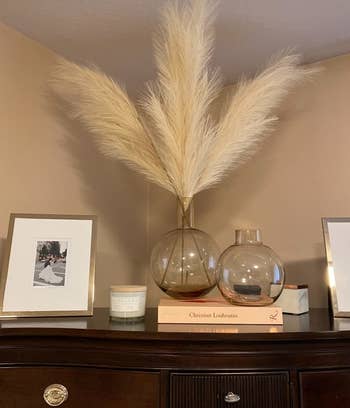 reviewer photo of the beige faux pampas plumes in a glass vase on top of a wooden cabinet