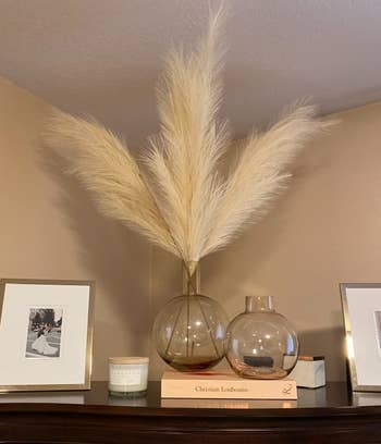 reviewer photo of the beige faux pampas plumes in a glass vase on top of a wooden cabinet