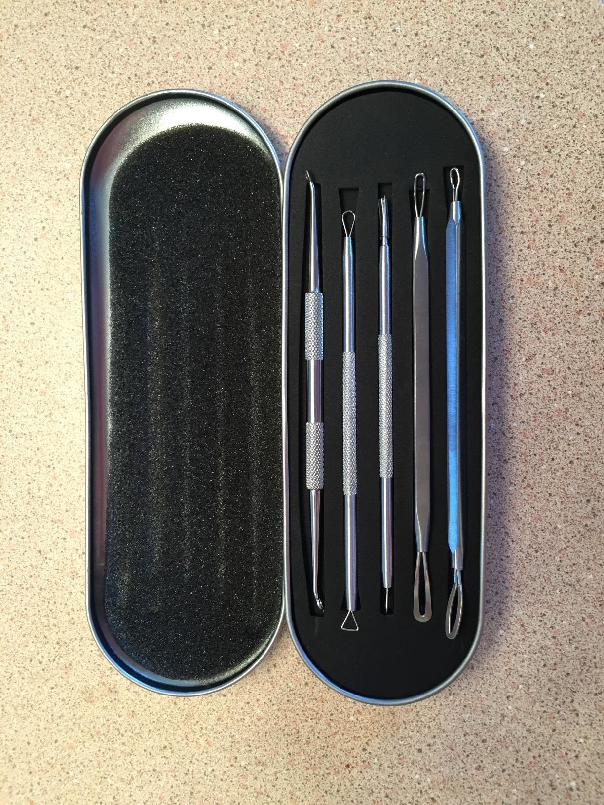 A silver tin (shaped like an eyeglass case) is black on the inside and has five skinny slots where the blackhead remover tools are sitting. A hand is holding one of the tools which has a looped end.