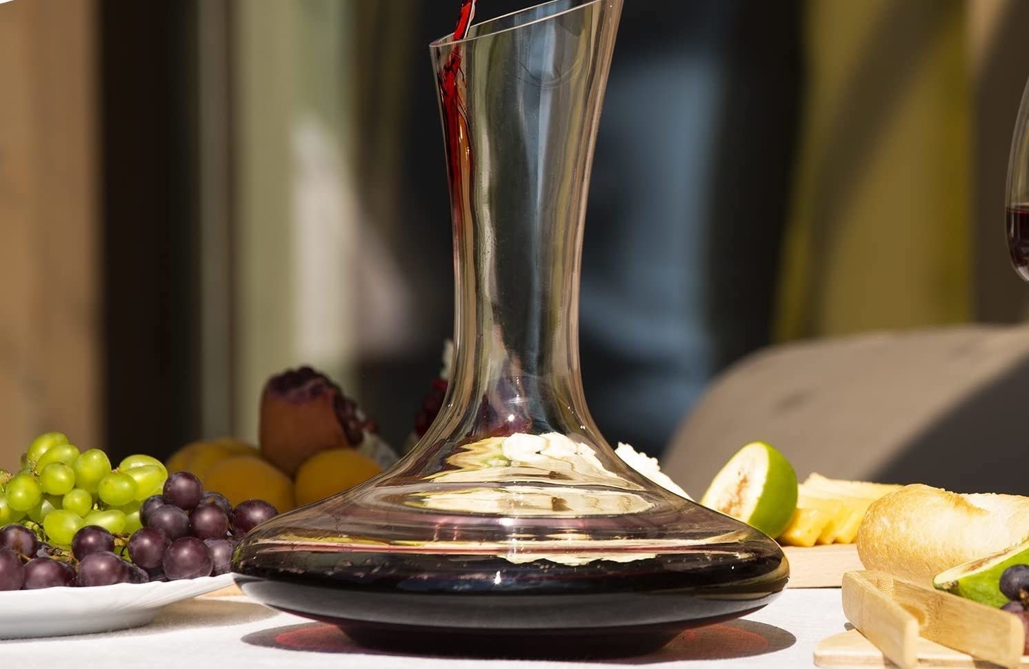 Wine being poured into a decanter on a table with fruit and crackers surrounding it