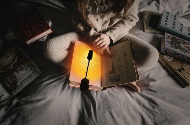 A person sitting on a bed reading a book with the amber light attached to the book