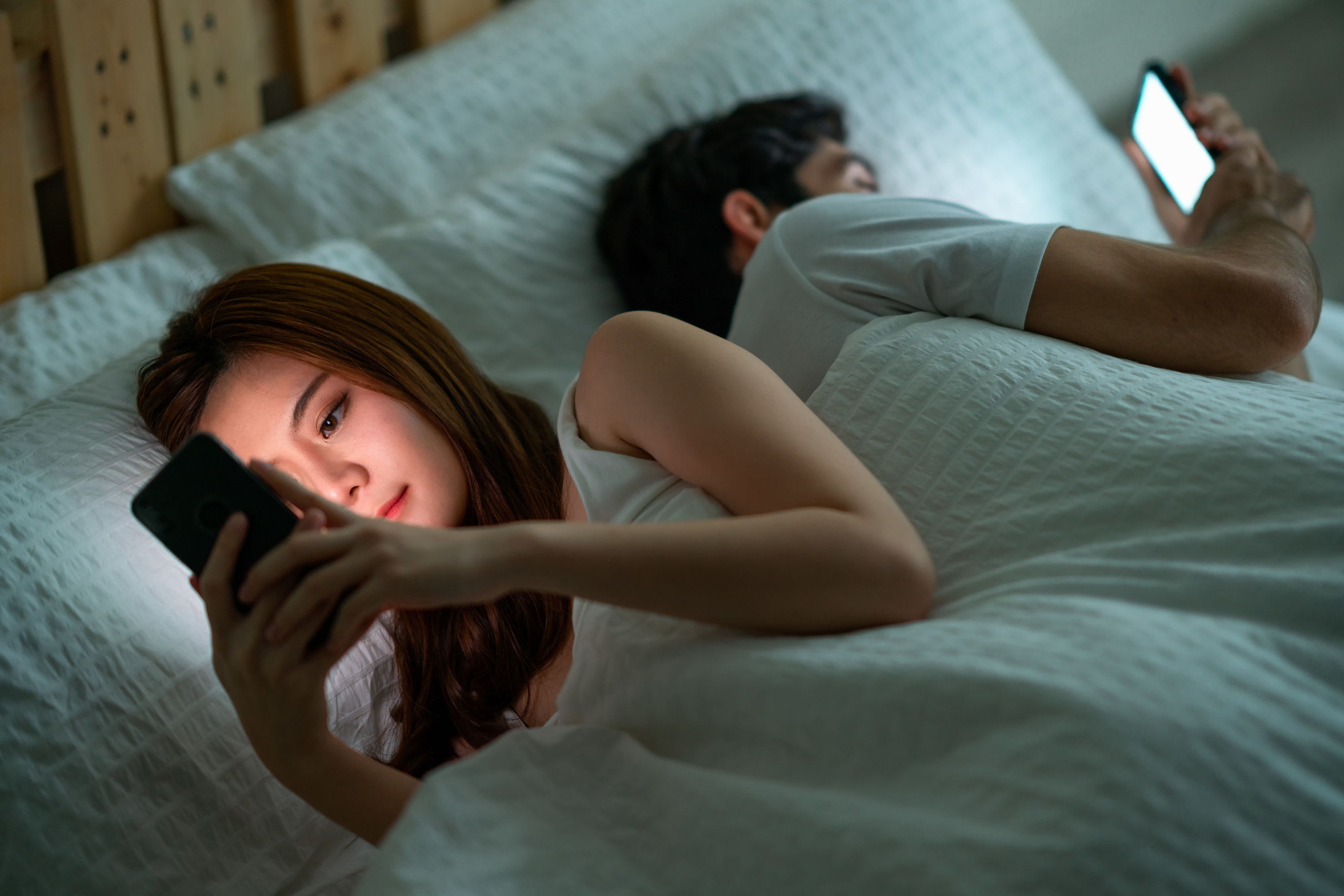 A man and a woman each on their phones in bed with their backs facing each other