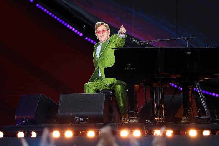 Elton wears a green suit and points to the audience from his piano