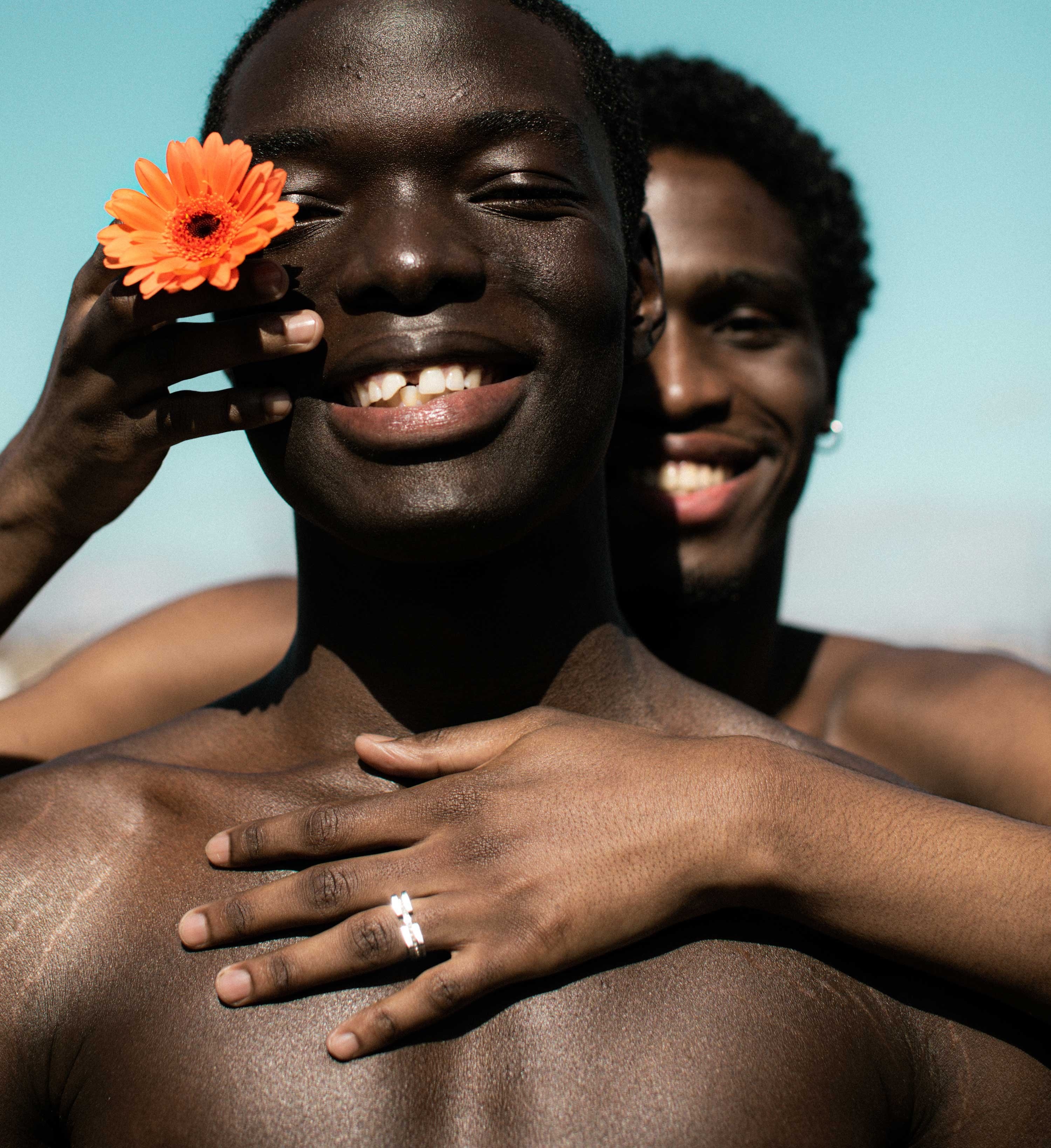 Two smiling people, with one standing behind the other and touching the other&#x27;s bare chest and holding a flower up to their eye