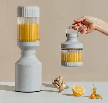 the gray blender filled with an orange smoothie and a hand holding a smaller blending vessel with an attached carry handle