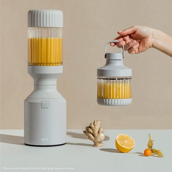 the gray blender filled with an orange smoothie and a hand holding a smaller blending vessel with an attached carry handle