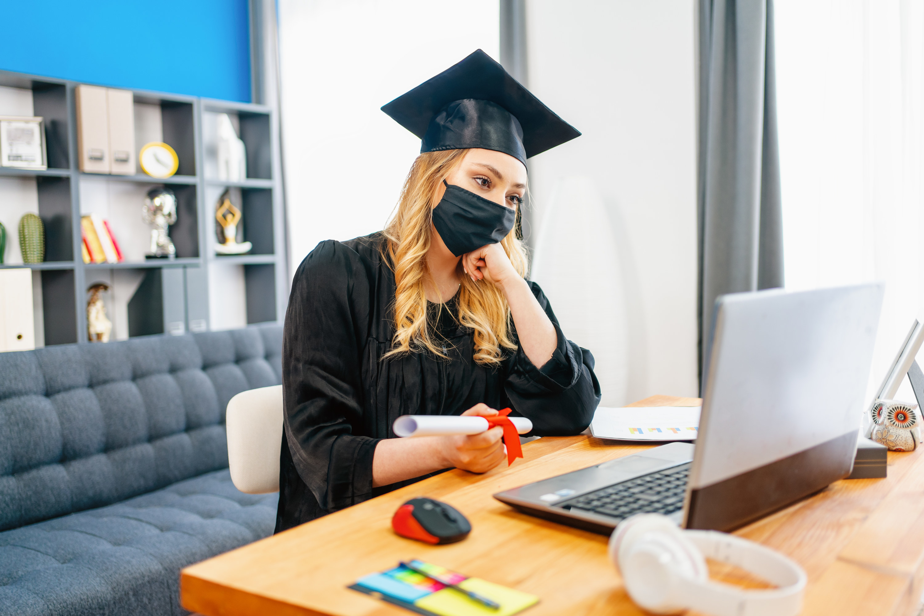 A girl in a graduation cap and gown sits in front of her computer for a virtual graduation ceremony