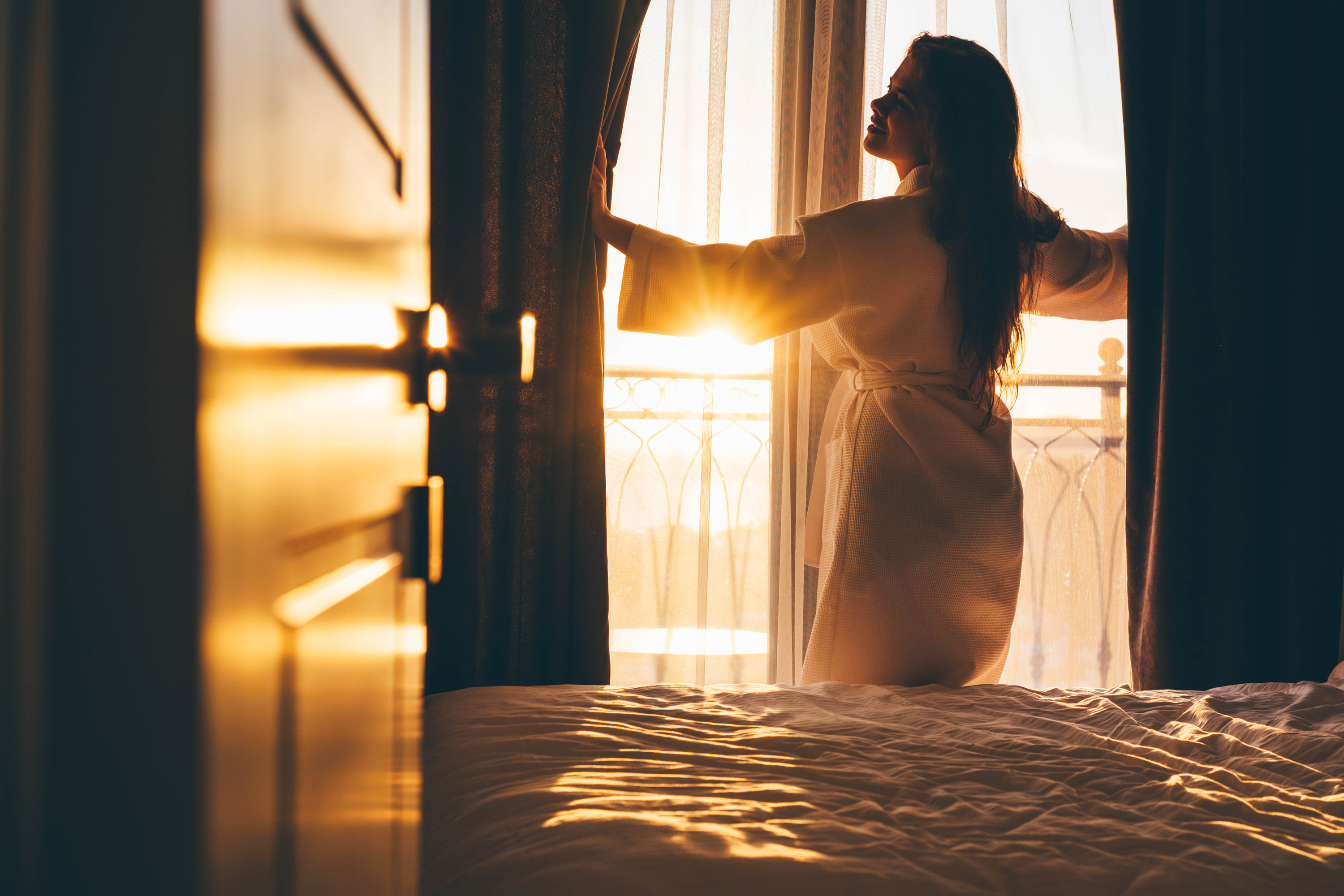 A woman in a bathrobe opens the curtains to a vibrant sunrise