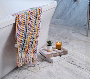 the rainbow colored towel draped over the side of a bathtub, with some books, a candle, and a plant on the floor nearby