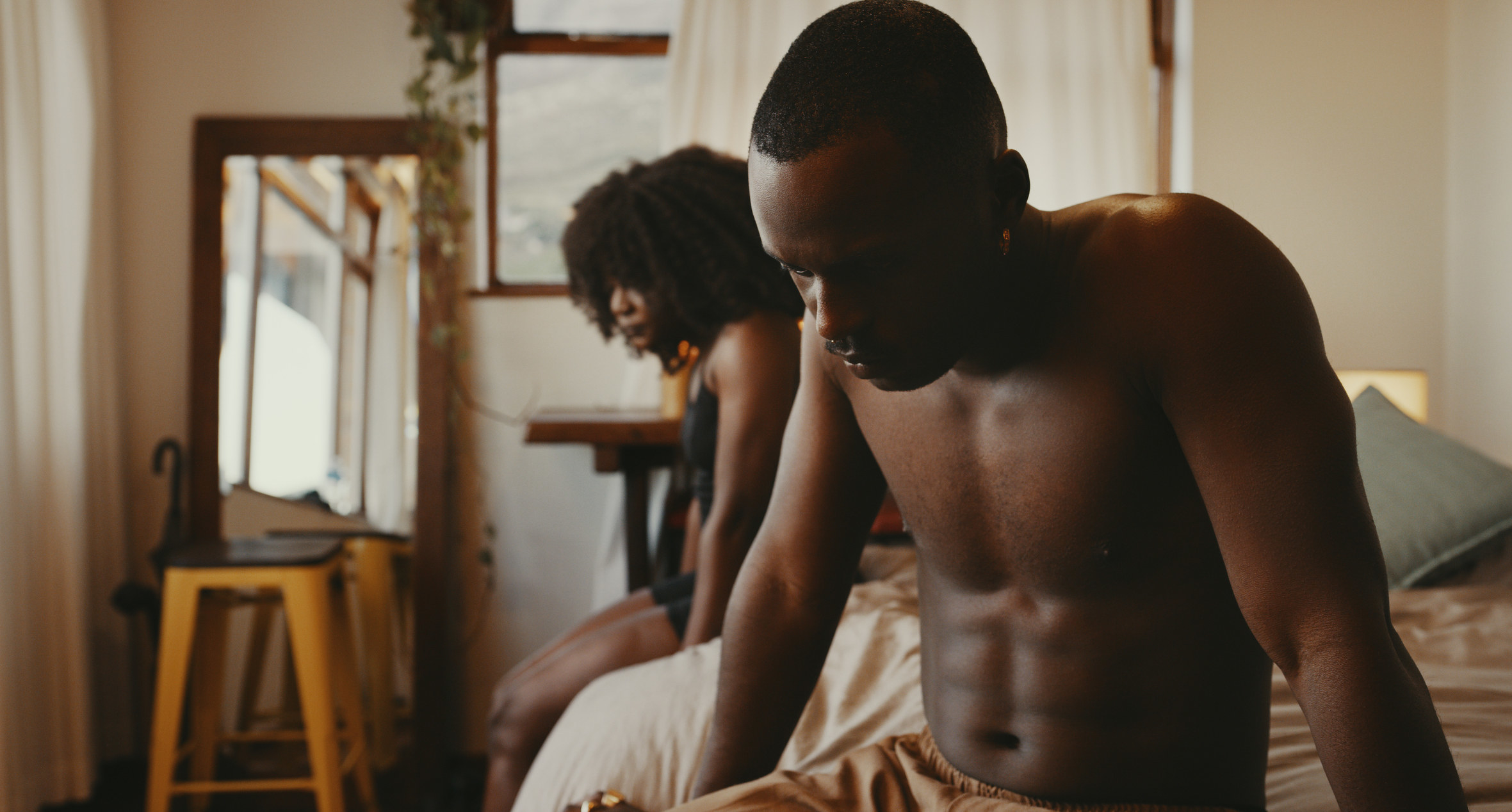 Shirtless man on bed sits far away from his partner, looking dejected