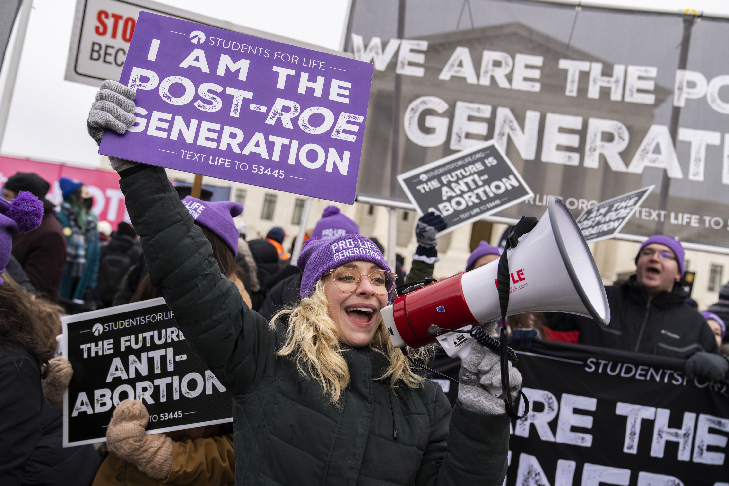 A woman wearing a beanie that reads &quot;pro-life generation&quot; talks into a megaphone and holds a sign reading &quot;I am the post-Roe generation&quot;