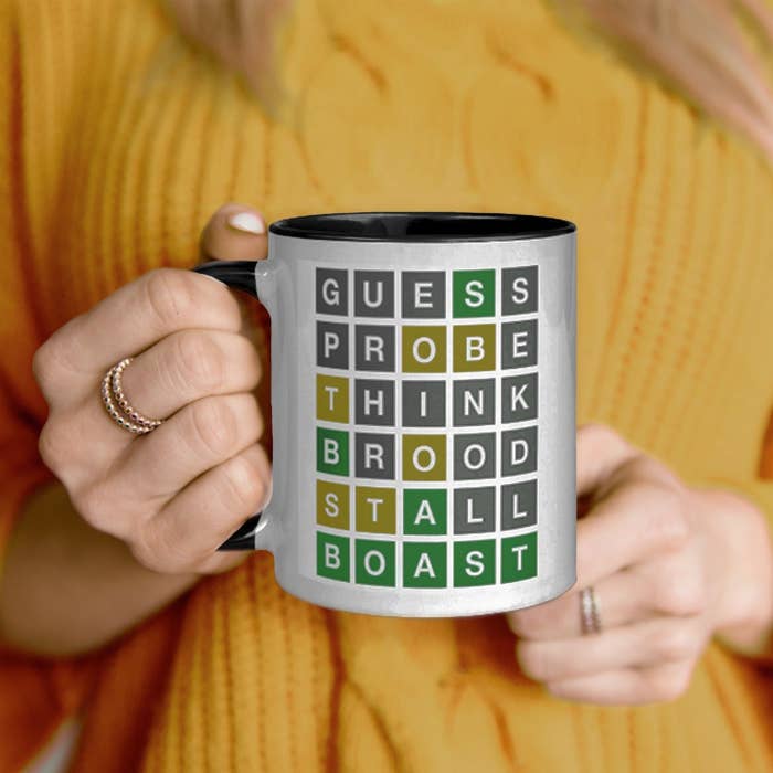 person holding up a mug with a wordle design on it showing the words &quot;guess, probe, think, brood, stall, boast&quot;