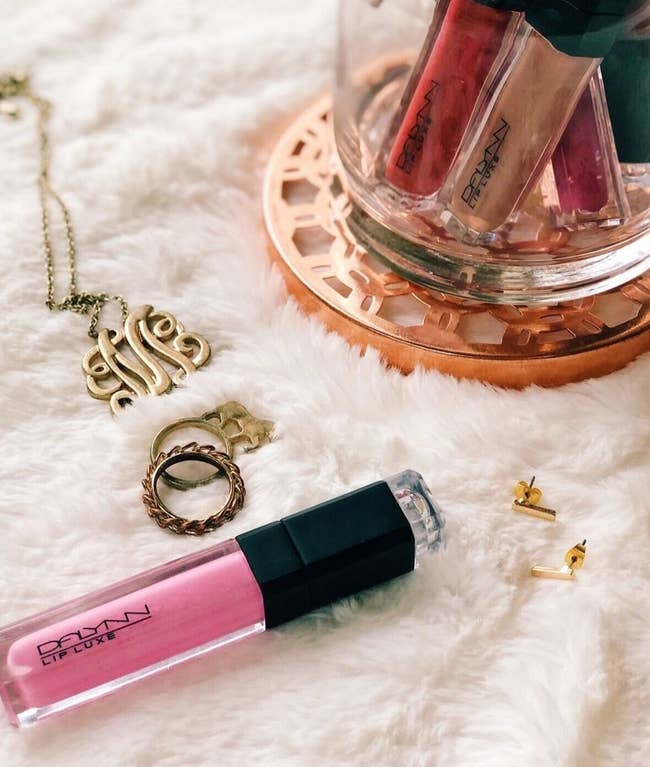 Pink and black tube of lip gloss next to jar of products in other shades and gold jewelry