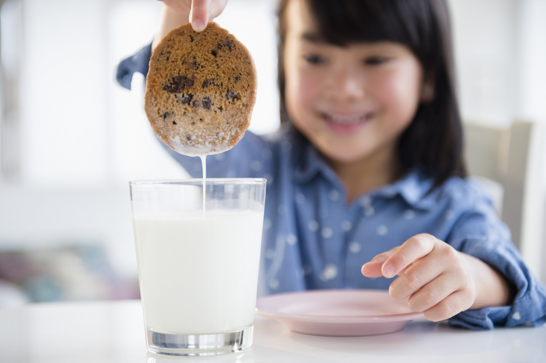 A photo of a girl at a dining table holding a cookie above a glass of milk she has just dunked in