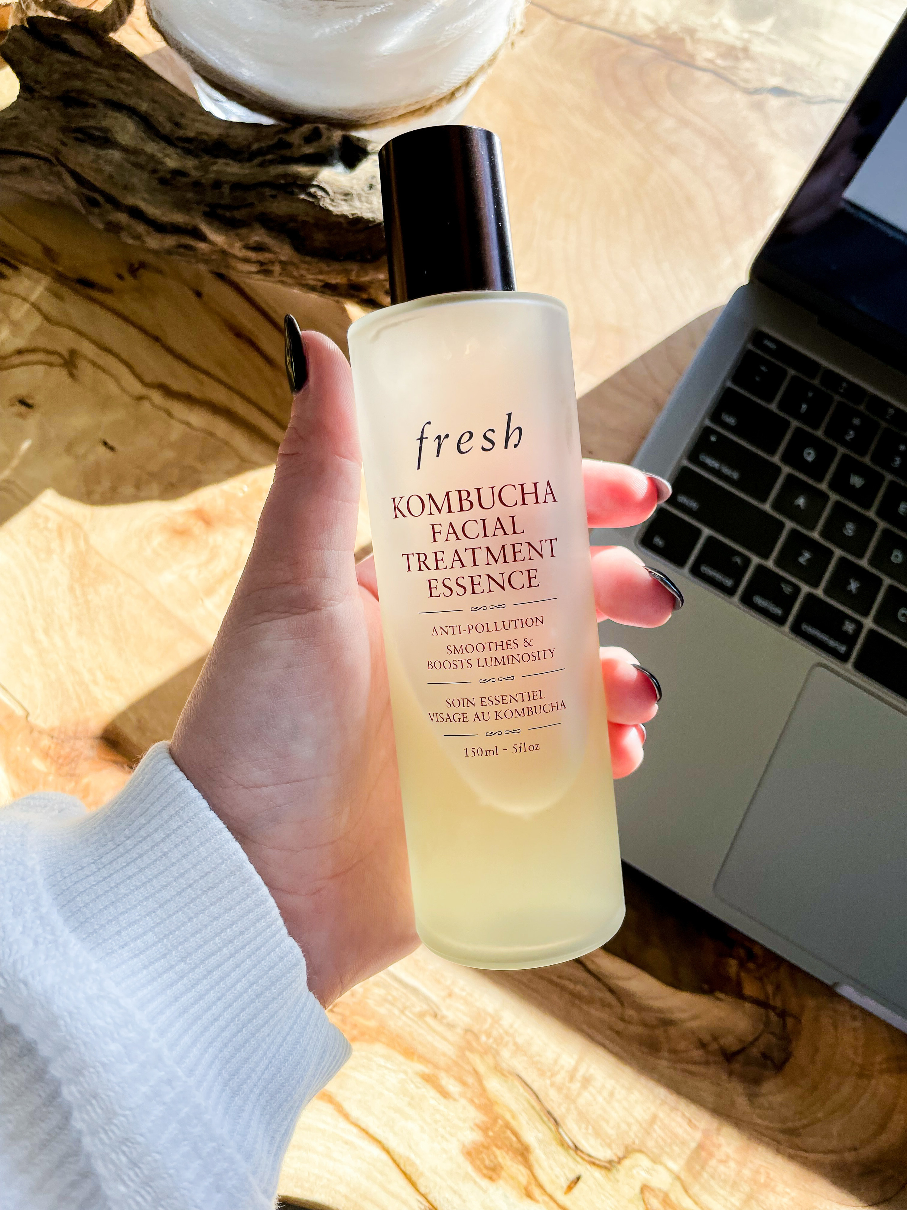 A person holding up a bottle of the face essence against a tabletop and laptop