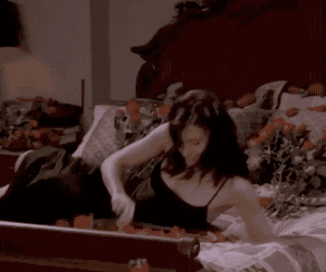 Courtney Cox as Monica Geller lays on a bed with roses on it and tries to put a rose in her mouth before a thorn stabs her and she spits it out in &quot;Friends&quot;