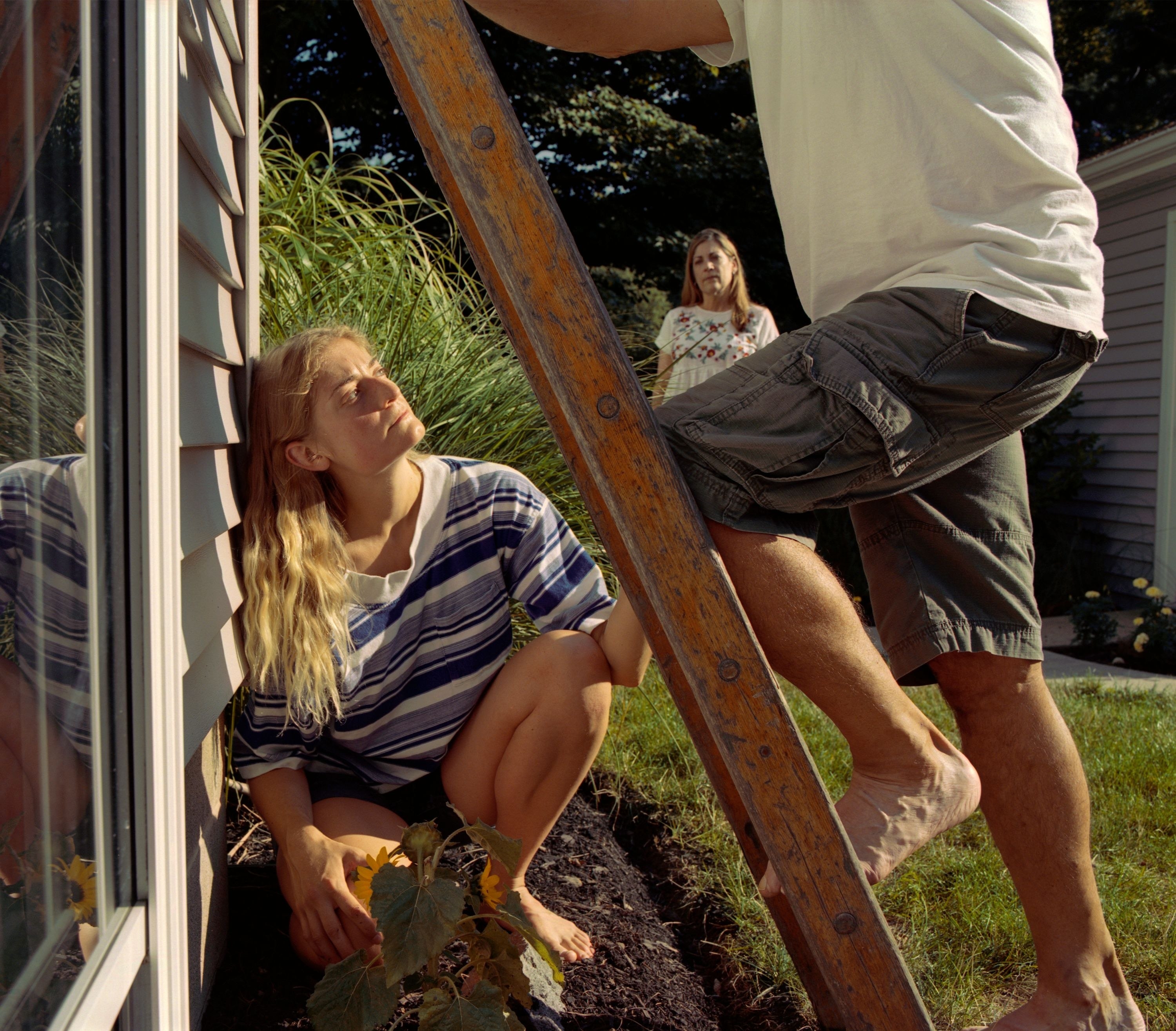 A barefoot woman kneels in the soil by the side of a house and looks up at a barefoot man climbing a ladder up against the house