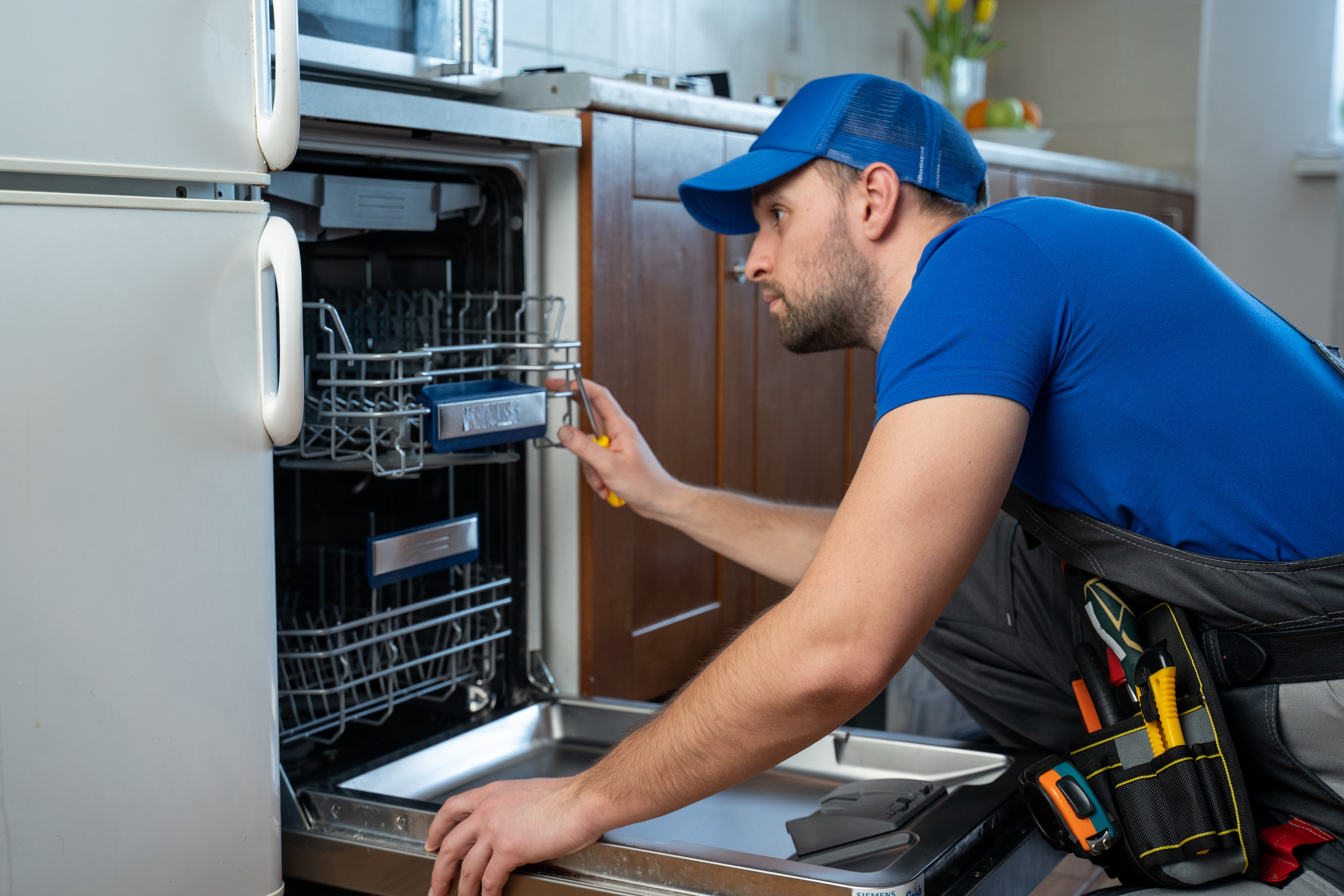A repairman wearing a blue shirt and hat looking into someone&#x27;s dishwasher