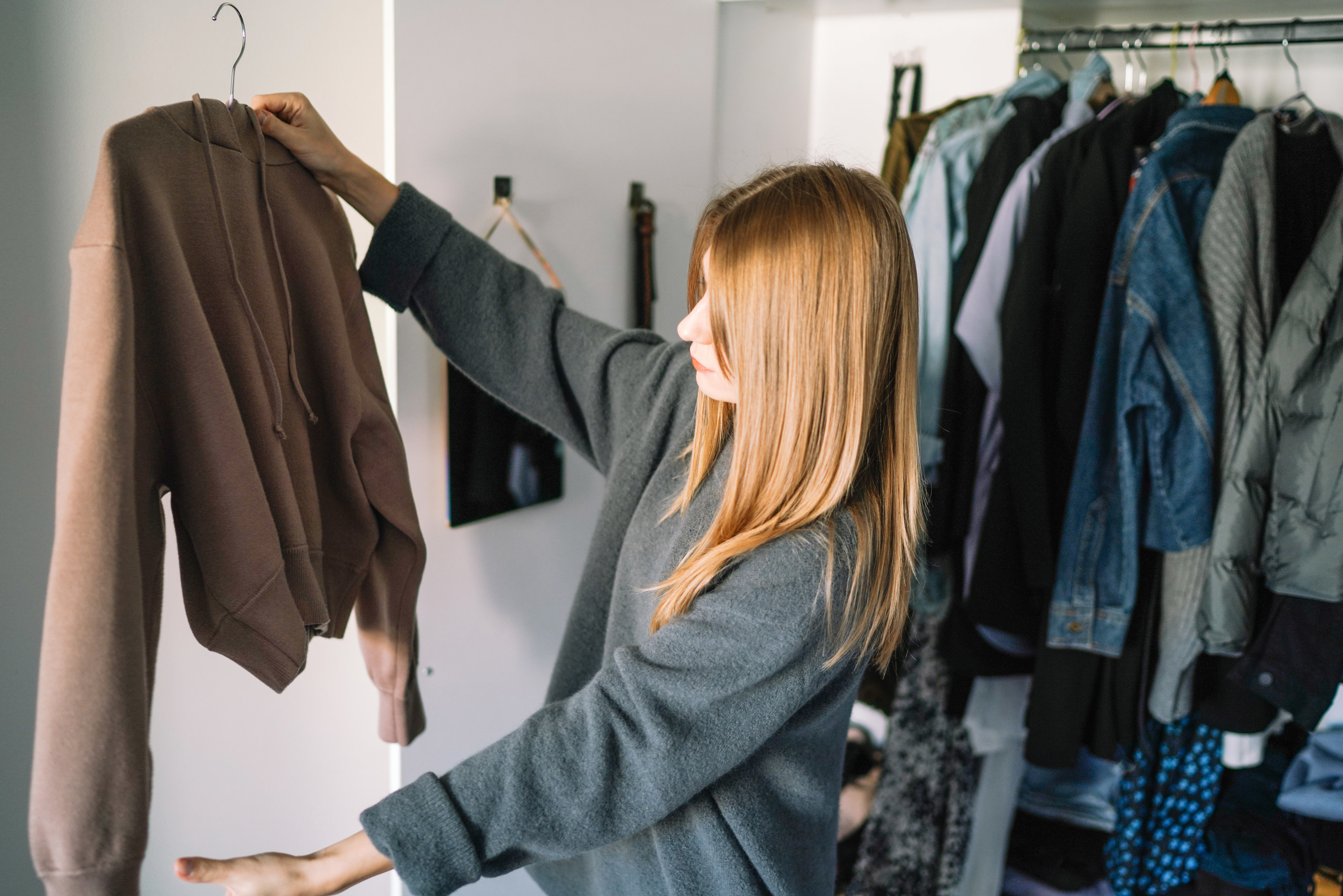 A woman standing in her closet holding up a brown hoodie on a hanger