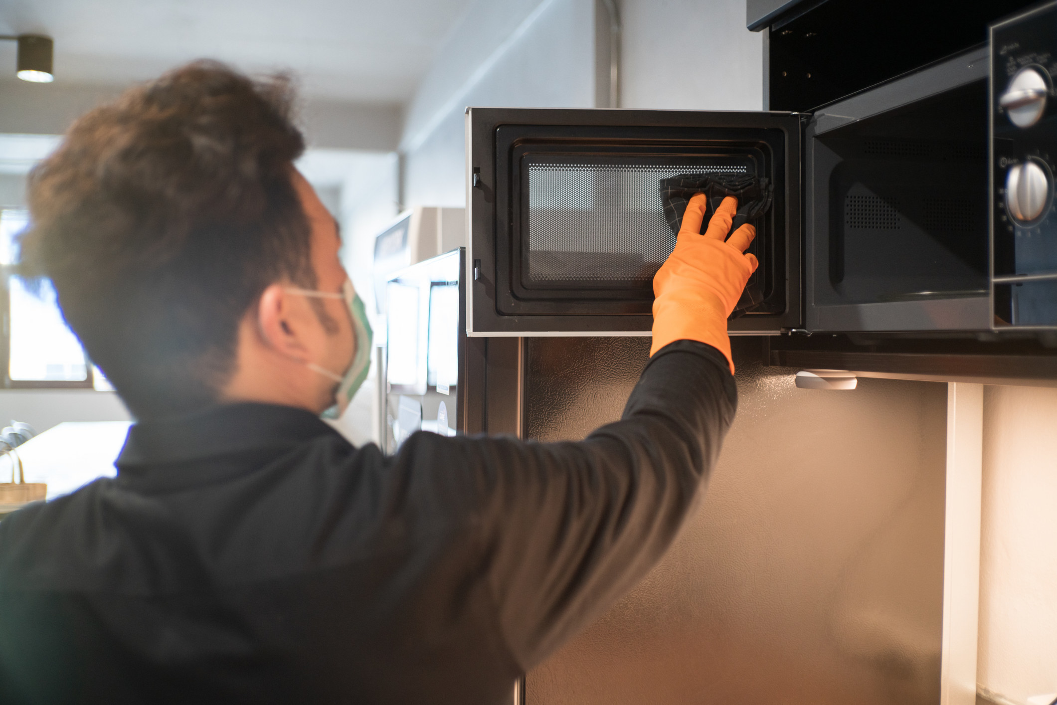 A man wearing a face mask wiping down the inside of a microwave with a towel