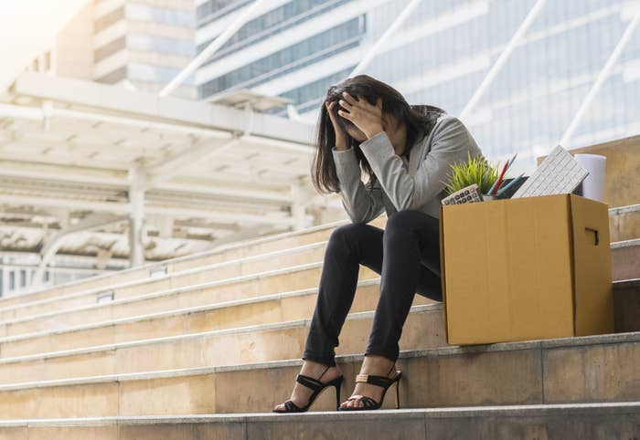 A woman sitting on the steps of an office building next to a cardboard box filled her belongings with her head in her hands