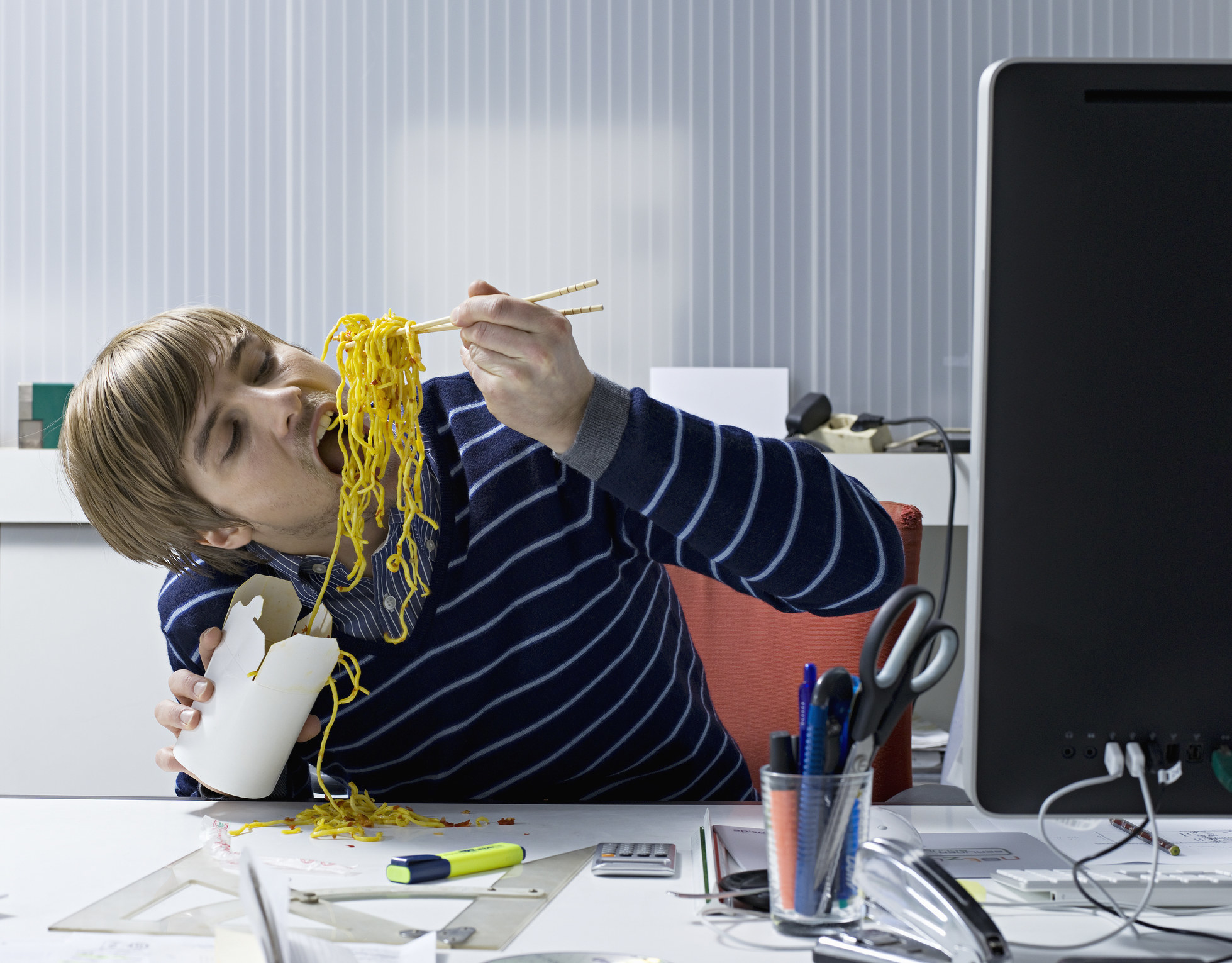 A person sitting at their work desk stuffing their face with take-out noodles