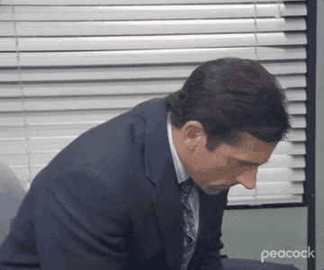 Steve Carell as Michael Scott sits at his desk looking at the ground before looking up suddenly and then turning to the camera in