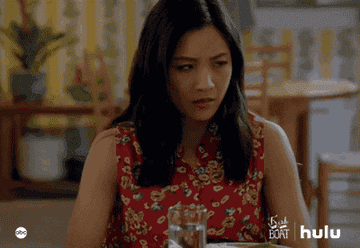 Constance Wu as Jessica Huang sits at a table and braces her elbows against it as she closes her eyes and lays her head down in her hands in