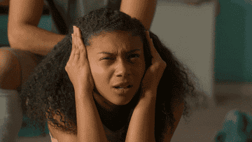 Sierra Capri as Monsé Finnie holds her hands over the sides of her head before raising them out and squinting upwards in