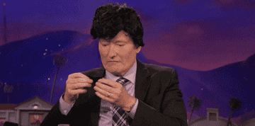 Conan O'Brien wears a short haired wig and places a thick fake mustache over his lip in