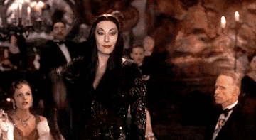 Anjelica Huston as Morticia Addams throws a knife at Raul Julia as Gomez Addams who catches it in his teeth and smiles in