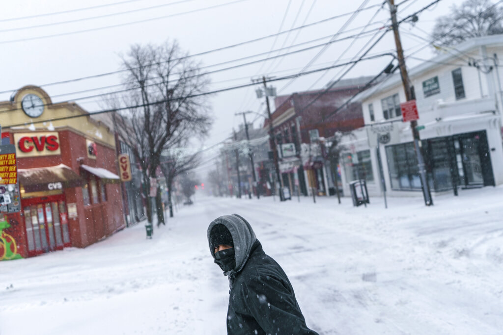 A person wearing a jacket with a hood and a face covering walks along an otherwise empty city intersection