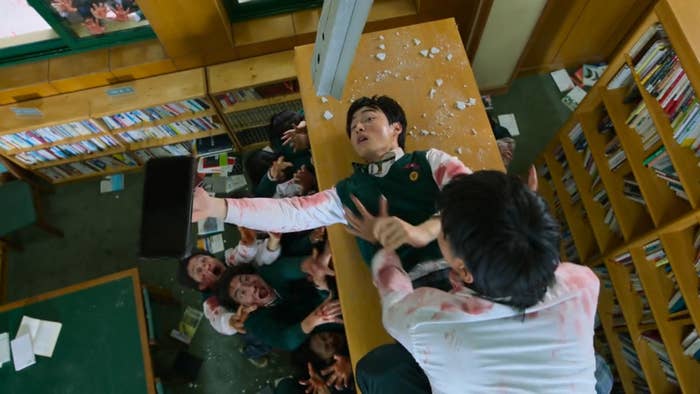 Two boys fight on the top of a bookshelf in a library full of zombies