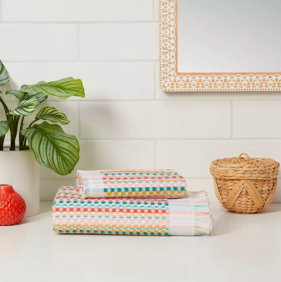 Folded towels sitting next to a plant and a basket in front of a tiled wall with a mirror on it