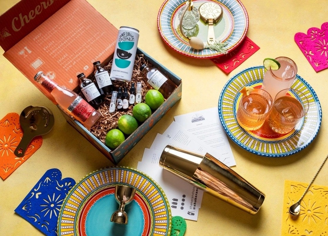 a cocktail kit filled with ingredient bottles and limes, surrounded by cocktail-making accessories