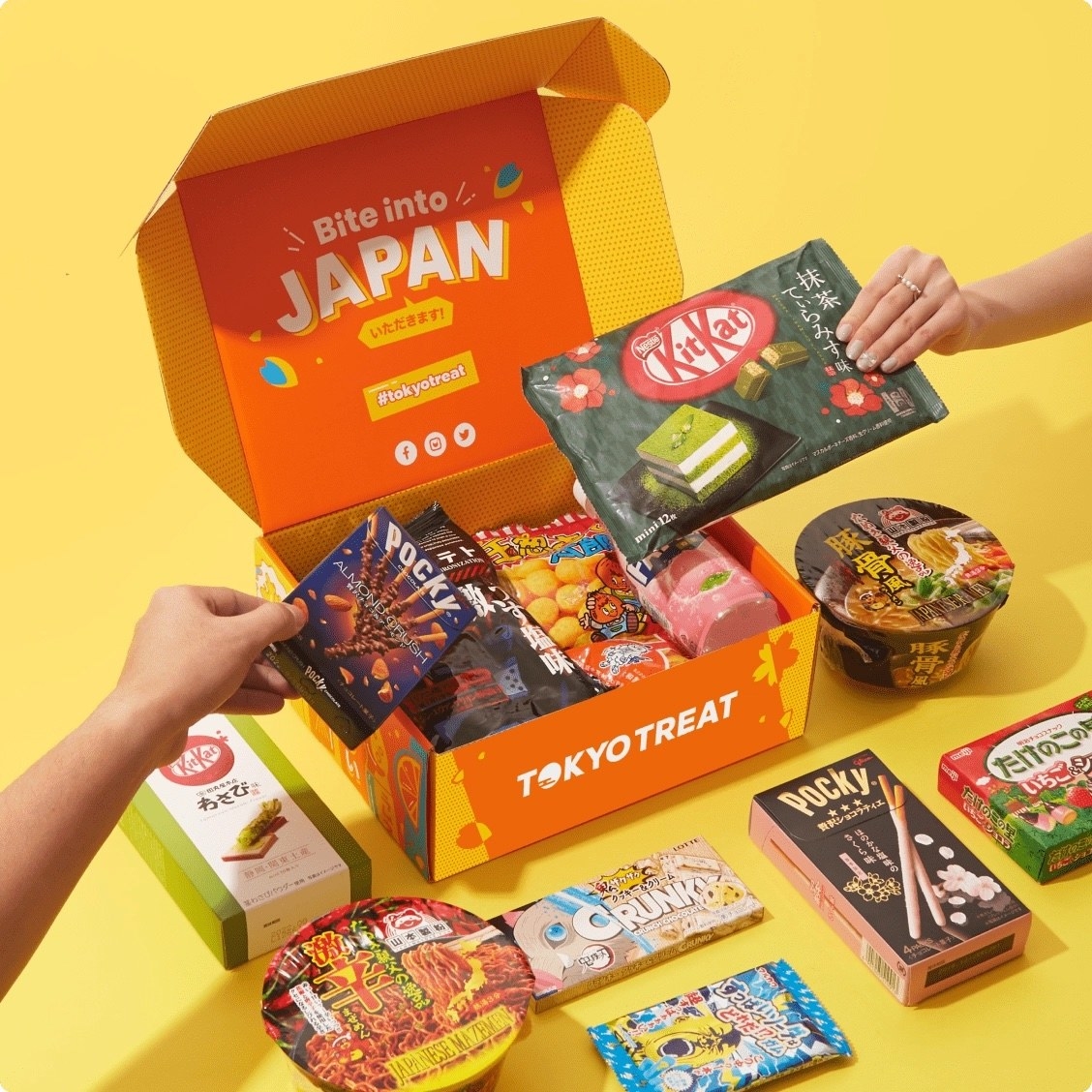 A Tokyo Treat box filled with assorted snacks, with hands holding packages of Pocky and KitKats