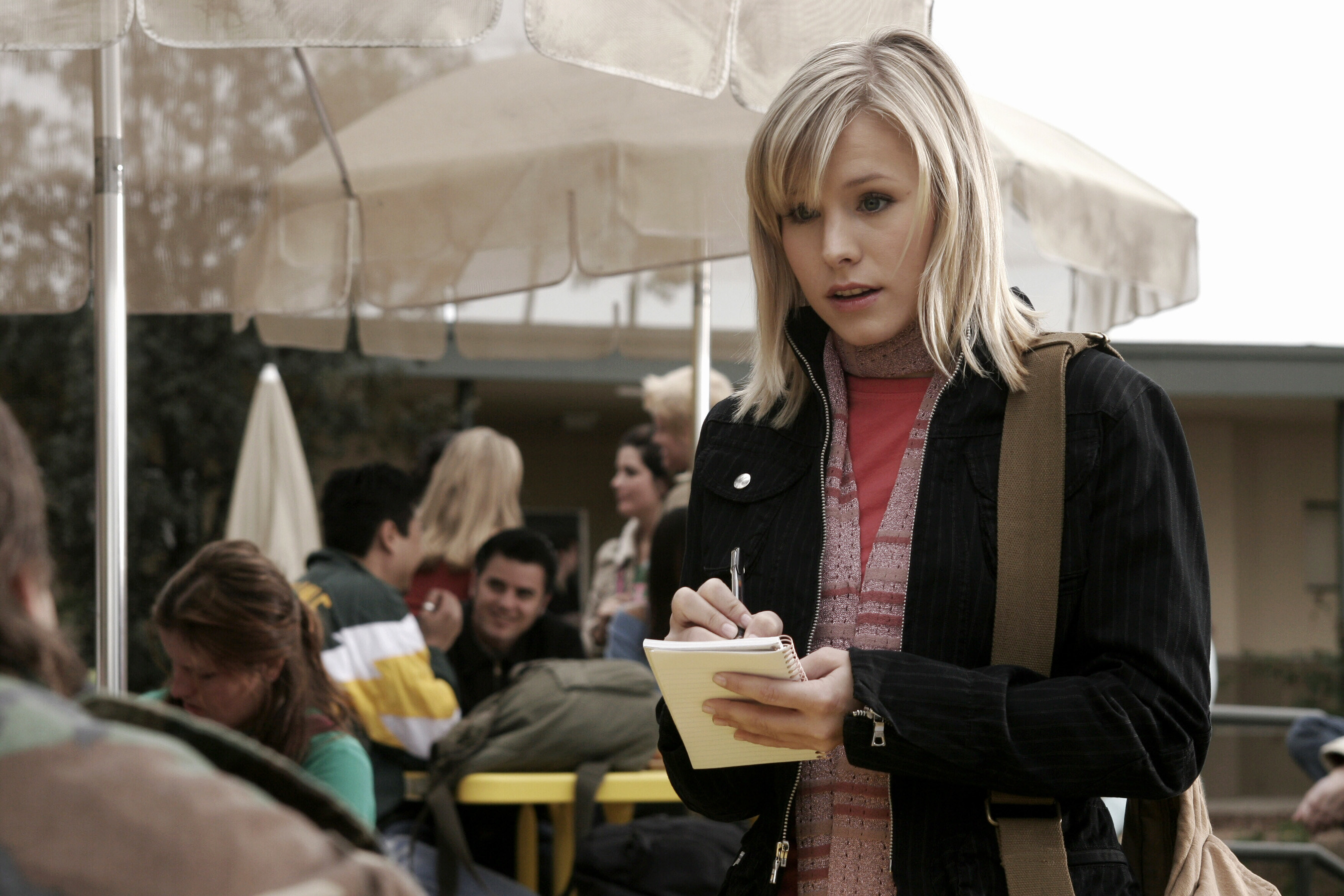 Veronica Mars writing in a notebook as she talks to someone sitting at a table