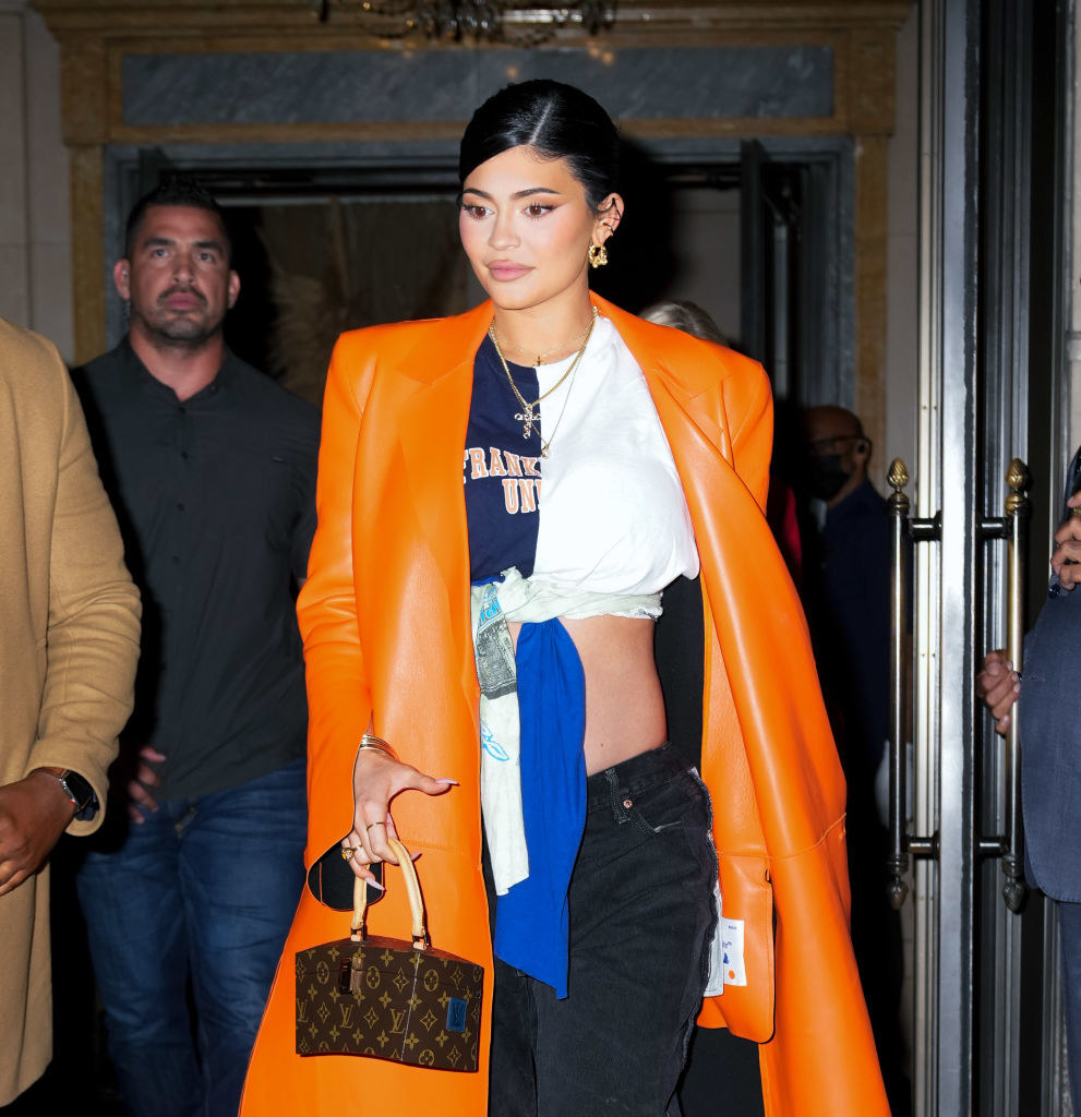 Kylie walking out of a building while wearing a long leather coat, jeans, and holding a small Louis Vuitton purse
