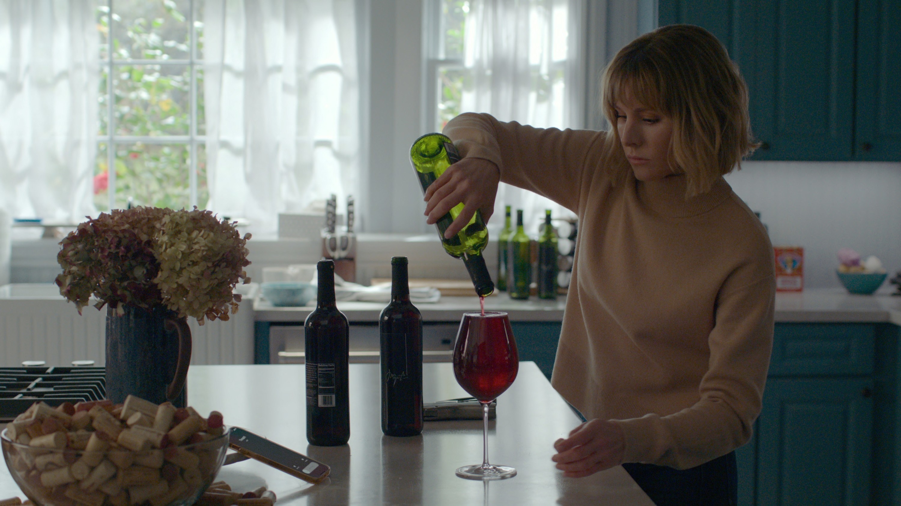 Kristin&#x27;s character standing at the kitchen island filling a wine glass to the brim and three empty wine bottles on the counter behind her