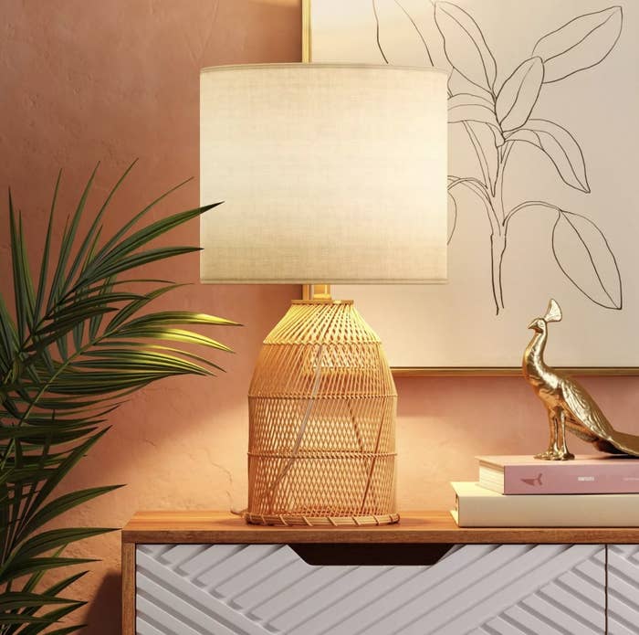 The light tan rattan lamp has a white linen shade with a warm orange glow coming from the bulb