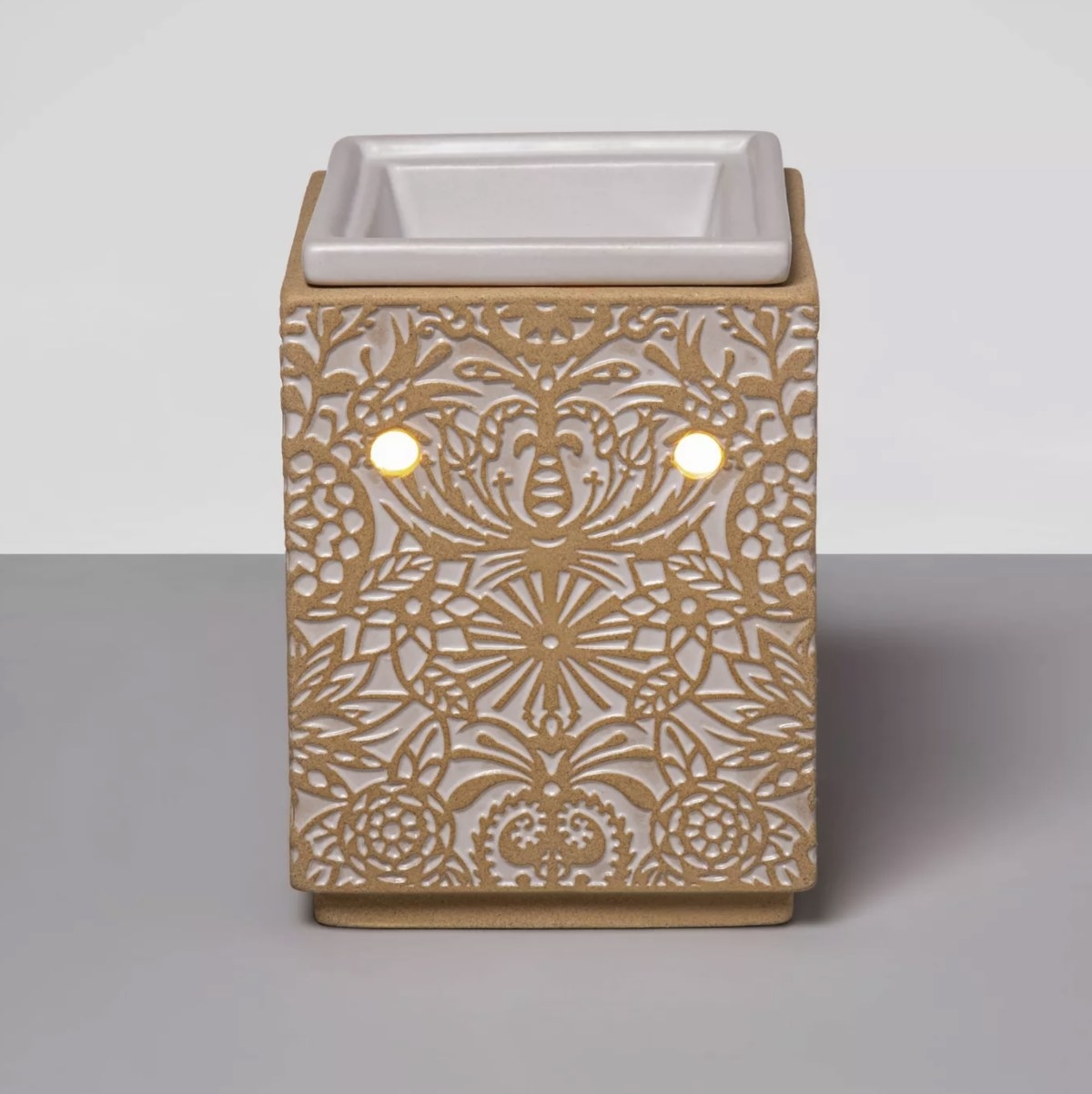 The tan and white embossed ceramic warmer has a white tray and two light holes glowing warm orange