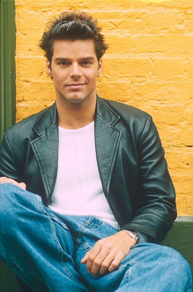 Ricky wearing blue jeans and a leather coat as he sits against a wall