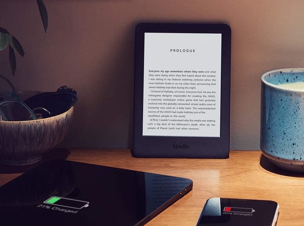 An Amazon kindle on a table surrounded by a tablet, a phone, a candle, and a plant