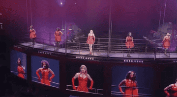 Taylor singing on stage with two background dancers and singers on both sides of her, as giant TVs project their image below them.