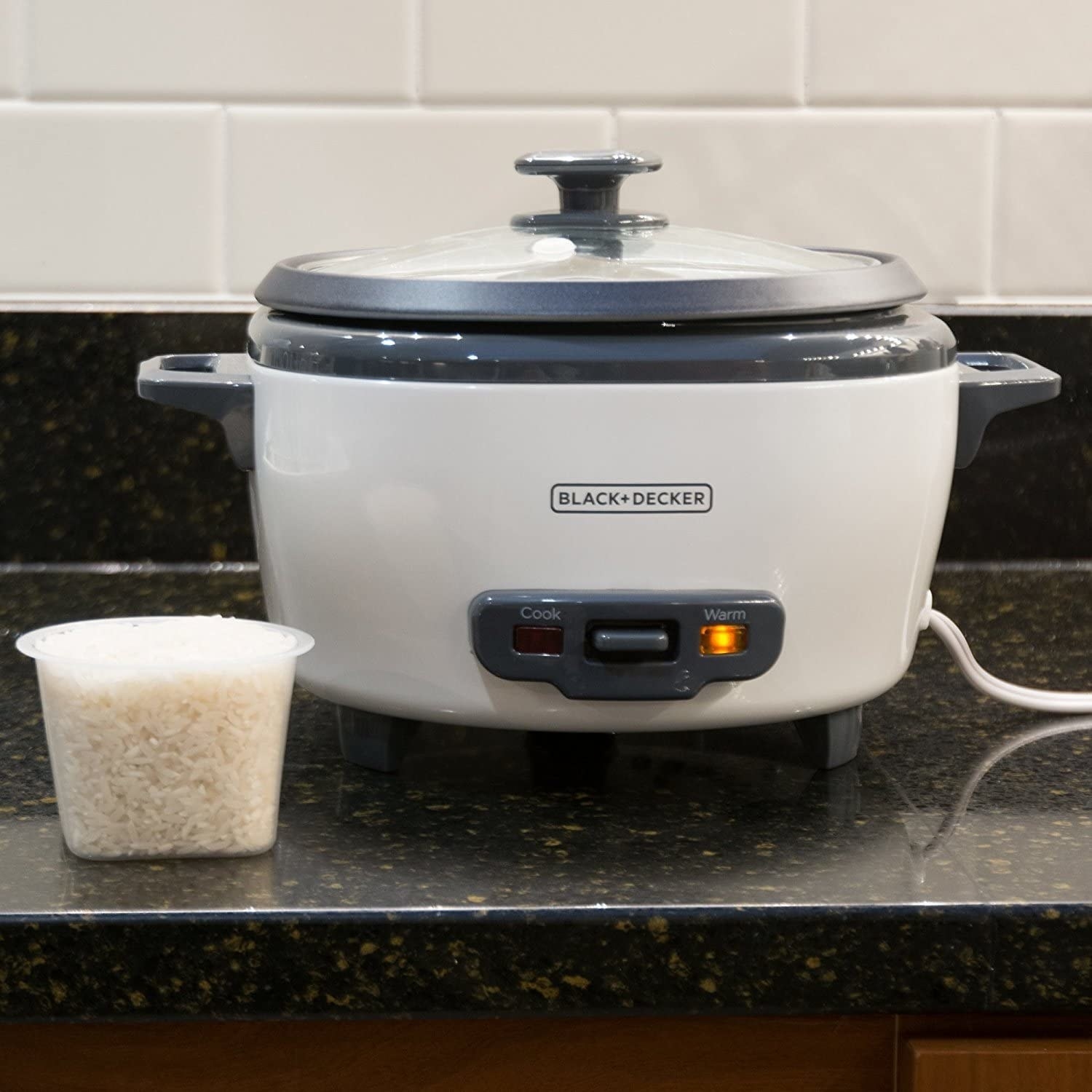 The rice cooker on a counter next to a container of rice