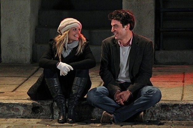 zoey and ted sit on a curb, wearing coats as if it&#x27;s cold, smiling at one another
