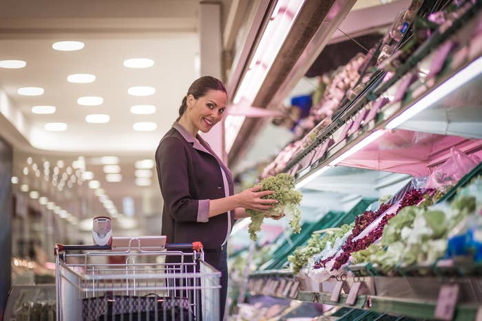 A woman smiling at the camera as she peruses the different lettuces at the grocery store