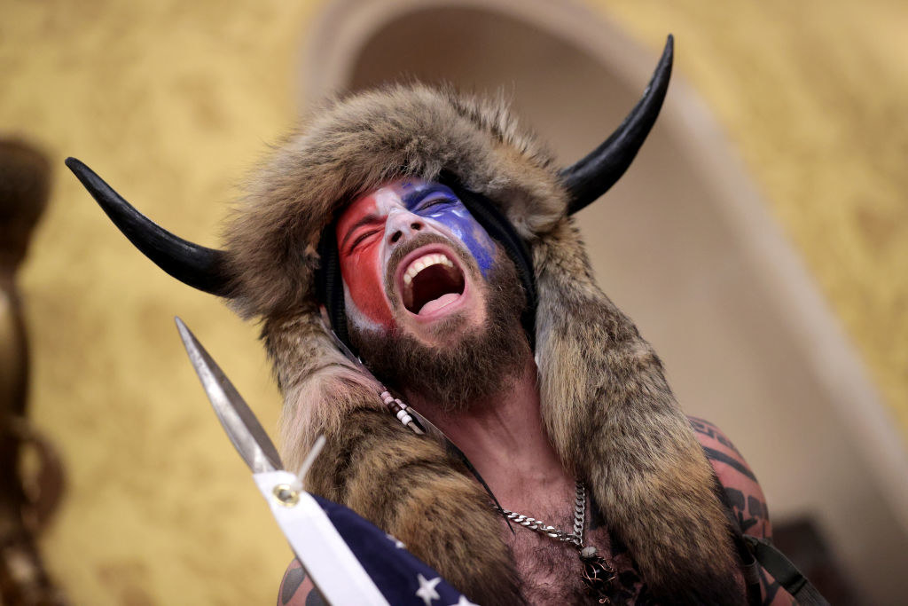Jacob Chansley wearing a horned Viking-esque fur hat and red, white, and blue face mask as he lets out a yell