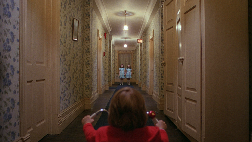 A boy riding a tricycle down a long hallway leading to twin girls.