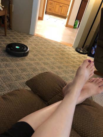 reviewer sits on couch while Roomba robot vacuum cleans their floor